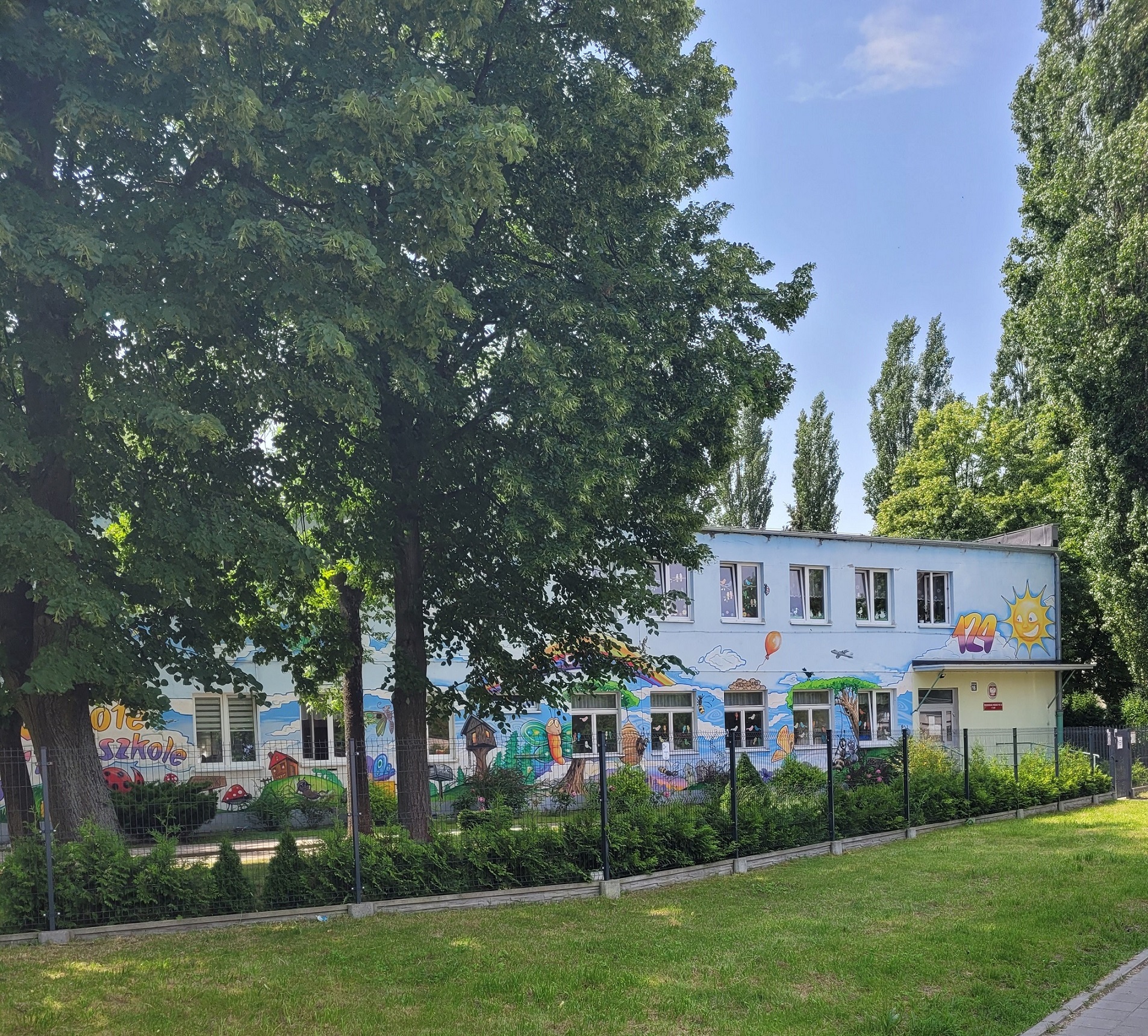 Zdjęcie budynku przedszkola pośród drzew. Budynek jest pomalowany na kolorowo i ogrodzony siatką.